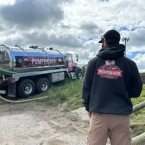 A person wearing a black hoodie with the PumperJack Septic logo stands outdoors facing a PumperJack Septic vacuum truck. The truck is silver with red accents and features the same company logo on its side. The scene is set under a partly cloudy sky, with hoses extending from the truck for septic service work.