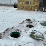 Snow-covered yard with open septic tank access lids showing winter septic service and cold-weather maintenance conditions
