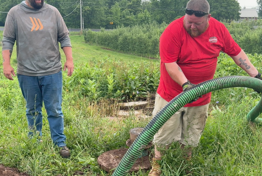 Two men performing septic pumping service in a grassy area, one man in a red shirt and gloves handling a green hose, while the other man in a gray shirt looks on, illustrating septic maintenance and compliance.